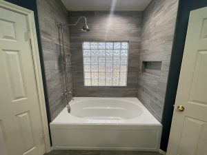Modern bathroom with a white bathtub, grey tiled walls, glass block window, wall-mounted showerhead, and a built-in niche. Doors are on both sides of the bathtub.
