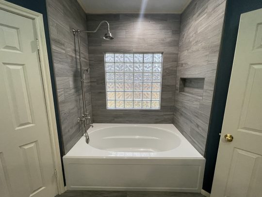 Modern bathroom with a white bathtub, grey tiled walls, glass block window, wall-mounted showerhead, and a built-in niche. Doors are on both sides of the bathtub.