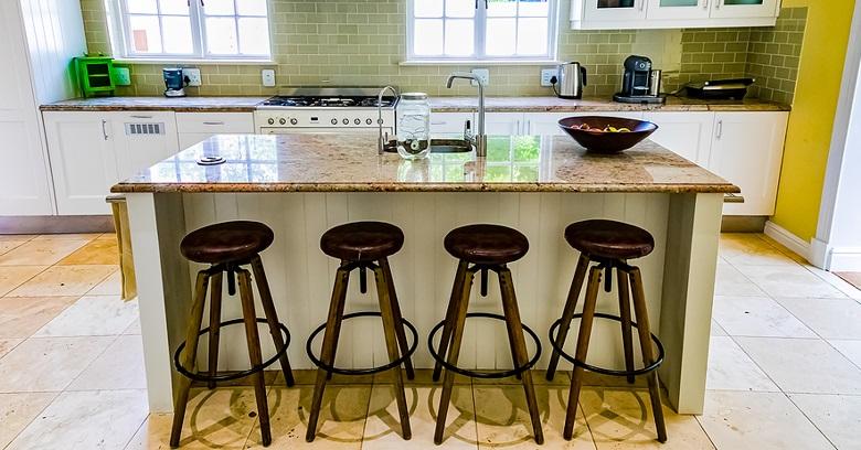 A modern kitchen with a central island, four bar stools, granite countertops, white cabinets, and a bowl of fruit on the island.