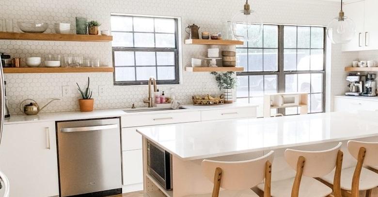 Modern kitchen with white cabinets, open wooden shelves holding dishes, large island with four chairs, stainless steel appliances, and large windows letting in natural light.