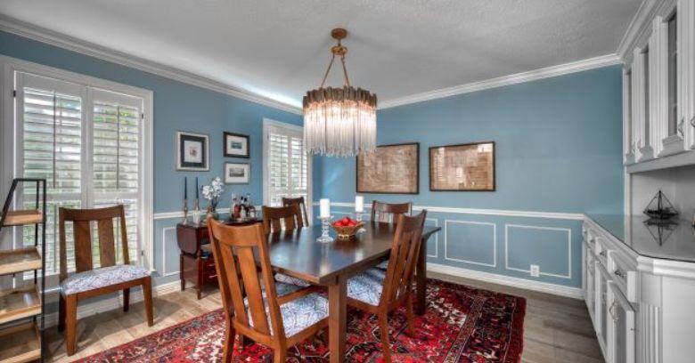 A dining room with a dark wood table, six chairs, a red patterned rug, blue walls with white trim, framed art, a chandelier, and built-in cabinets.