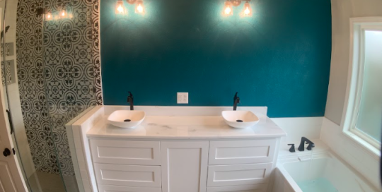 Modern bathroom with double sinks, teal accent wall, patterned shower tiles, white cabinetry, and a bathtub next to a large frosted window.