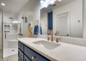 Modern bathroom with a white countertop, gold fixtures, blue cabinets, and a glass-enclosed shower with white tile. Blue towels and a gold decorative item are on the counter.