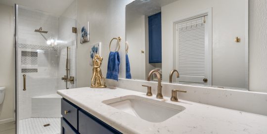 Modern bathroom with a white countertop, gold fixtures, blue cabinets, and a glass-enclosed shower with white tile. Blue towels and a gold decorative item are on the counter.