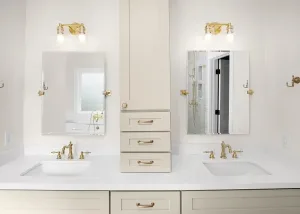 Modern bathroom with dual sinks, gold fixtures, two mirrors, white countertop, and a central beige cabinet with drawers and overhead lighting above each mirror.
