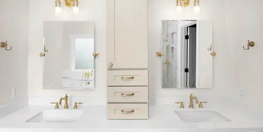 Modern bathroom with dual sinks, gold fixtures, two mirrors, white countertop, and a central beige cabinet with drawers and overhead lighting above each mirror.