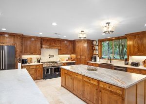 Spacious kitchen with wooden cabinets, stainless steel appliances, marble countertops, and a large island under ceiling lights; a window overlooks a green yard.