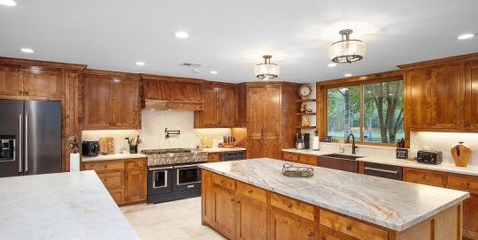 Spacious kitchen with wooden cabinets, stainless steel appliances, marble countertops, and a large island under ceiling lights; a window overlooks a green yard.