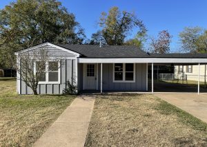 Single-story house with light blue siding, a covered carport, front door, and leafless shrub, set against a clear blue sky.