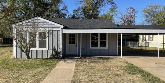 Single-story house with light blue siding, a covered carport, front door, and leafless shrub, set against a clear blue sky.