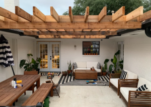 A covered outdoor patio with wooden pergola, dining table, and seating area, featuring black and white striped decor and potted plants.