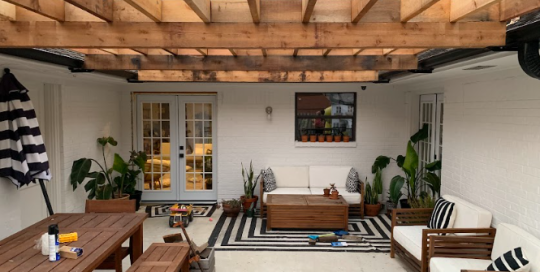 A covered outdoor patio with wooden pergola, dining table, and seating area, featuring black and white striped decor and potted plants.