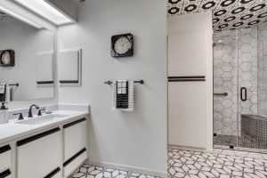 Modern bathroom with white and black decor, double sinks, wall clock, towel rack, geometric tile floor, and a glass shower enclosure.