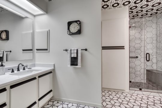 Modern bathroom with white and black decor, double sinks, wall clock, towel rack, geometric tile floor, and a glass shower enclosure.