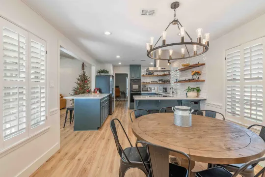 Bright kitchen and dining area with wooden floors, round dining table, black chairs, open shelves, modern light fixture, and Christmas decorations on the kitchen island in the background.