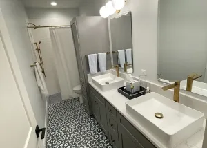 Modern bathroom with patterned tile floor, double sink vanity with gold fixtures, large mirrors, wall-mounted lights, and a shower with a white curtain in the background.