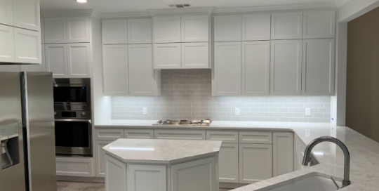 Modern kitchen with white cabinets, marble countertops, stainless steel appliances, and an island with a sink under recessed ceiling lights.