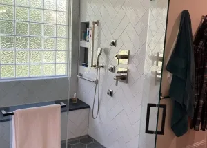 Modern bathroom shower with glass door, white herringbone tiles, built-in shelves, silver fixtures, bench with towel, and hexagonal tile flooring.