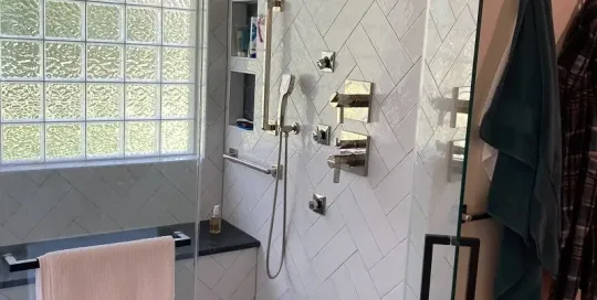 Modern bathroom shower with glass door, white herringbone tiles, built-in shelves, silver fixtures, bench with towel, and hexagonal tile flooring.