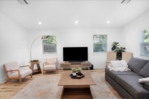 Modern living room with a gray sofa, two light armchairs, a wooden coffee table, TV on a wooden stand, floor lamp, potted plant, and two windows letting in natural light.