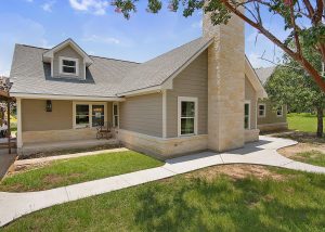 A beige house with a stone chimney, white trim, and a gray roof, surrounded by green grass, trees, and a curved concrete walkway.