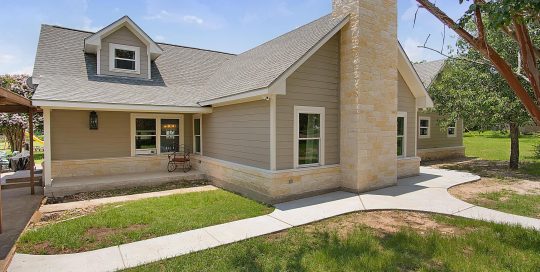 A beige house with a stone chimney, white trim, and a gray roof, surrounded by green grass, trees, and a curved concrete walkway.