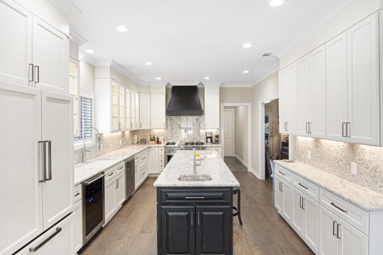 Modern kitchen with white cabinets, granite countertops, a center island with a black base, stainless steel appliances, and wood flooring. Bright lighting and ample workspace are visible.