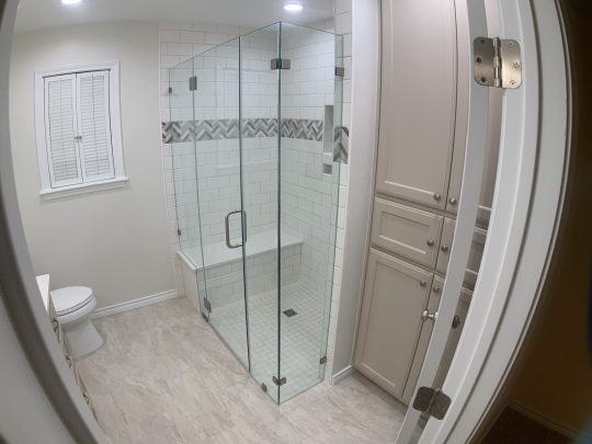 A modern bathroom with a glass-enclosed shower, beige tile floor, white cabinets, and a toilet near a window with shutters.